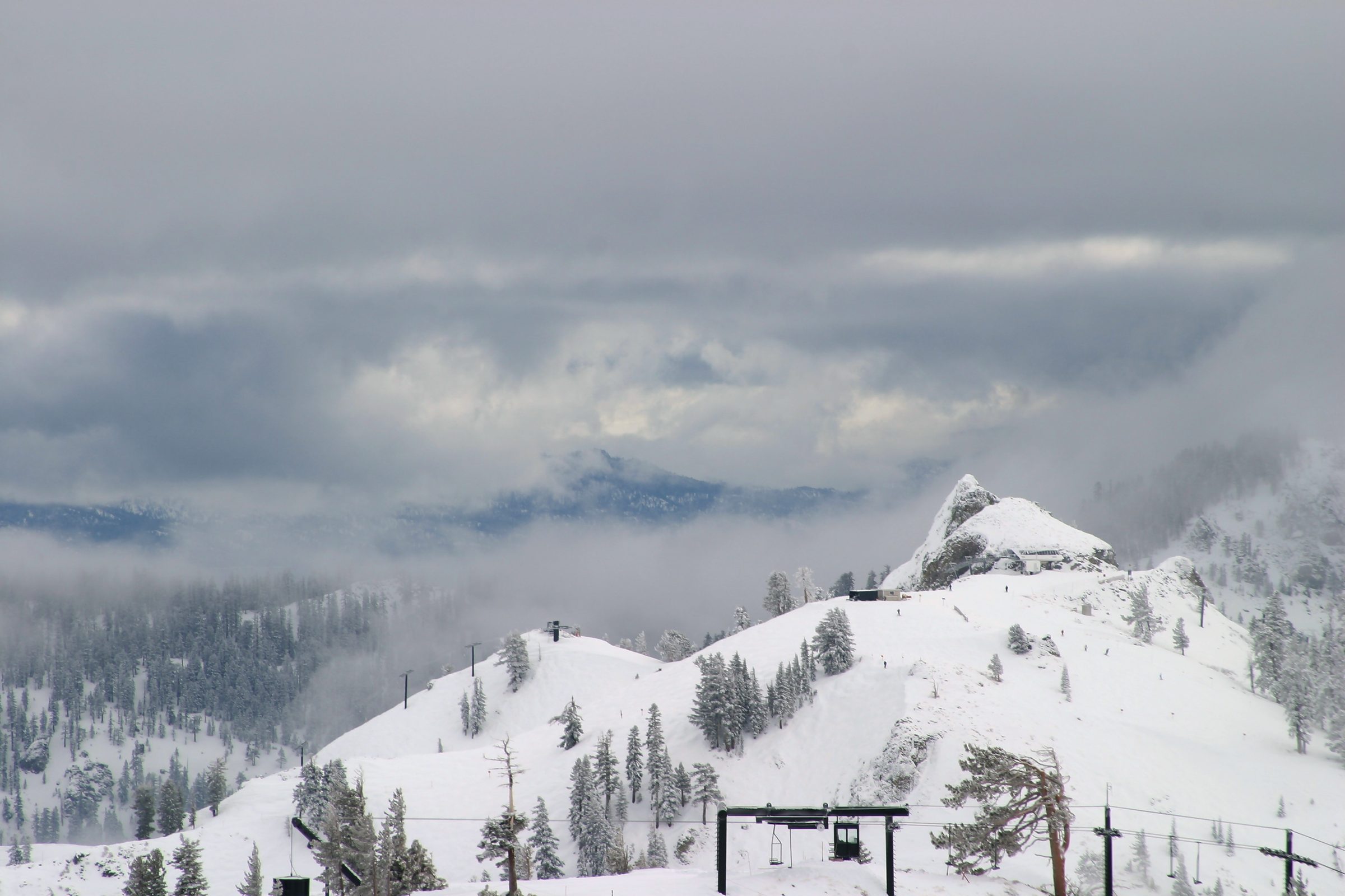 Snowy mountain peak rising from fog with faint ski-lift cables.