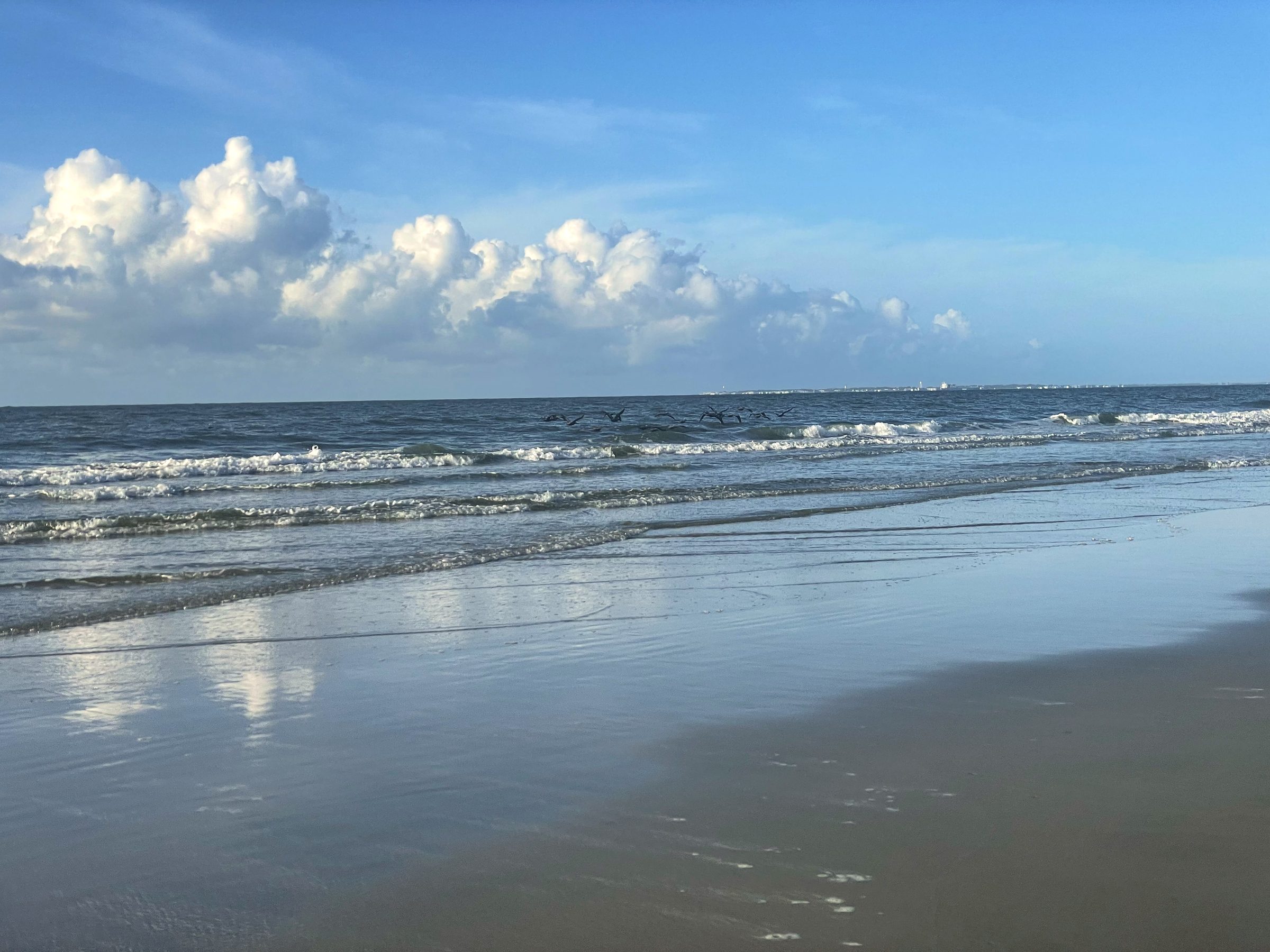 Gentle waves on flat sand under bright blue sky with clouds.