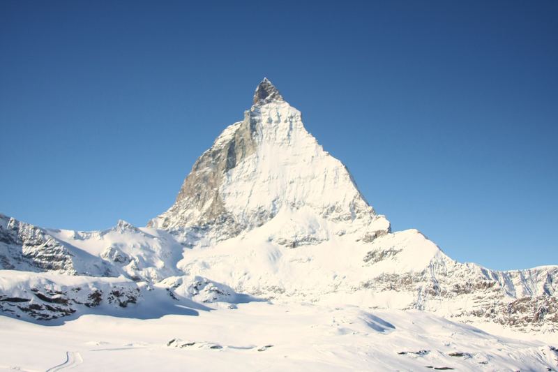Sunlit four-sided snowy summit with rocky base.