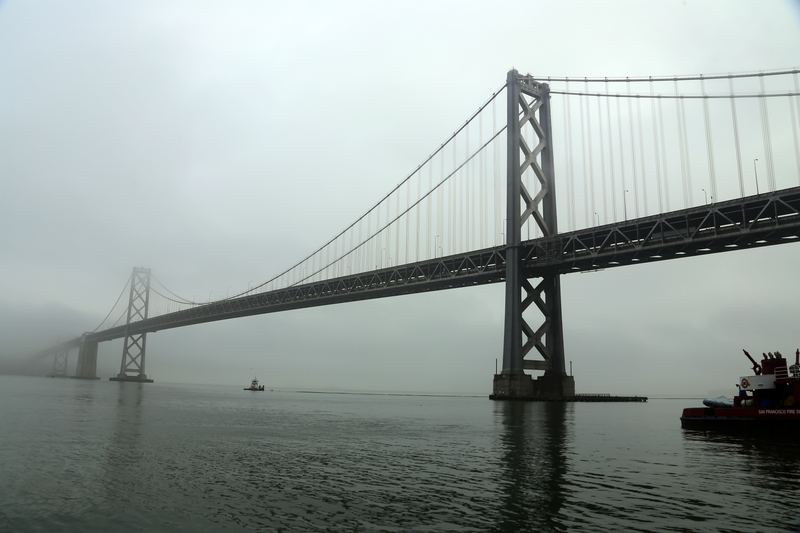 Bay Bridge X-bracing disappearing into fog over calm grey water.