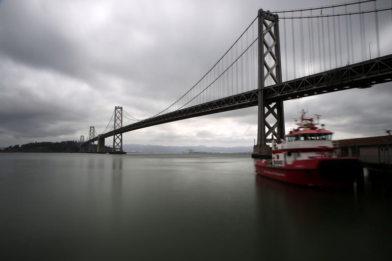 Low-angle Bay Bridge with a red fireboat under grey sky.