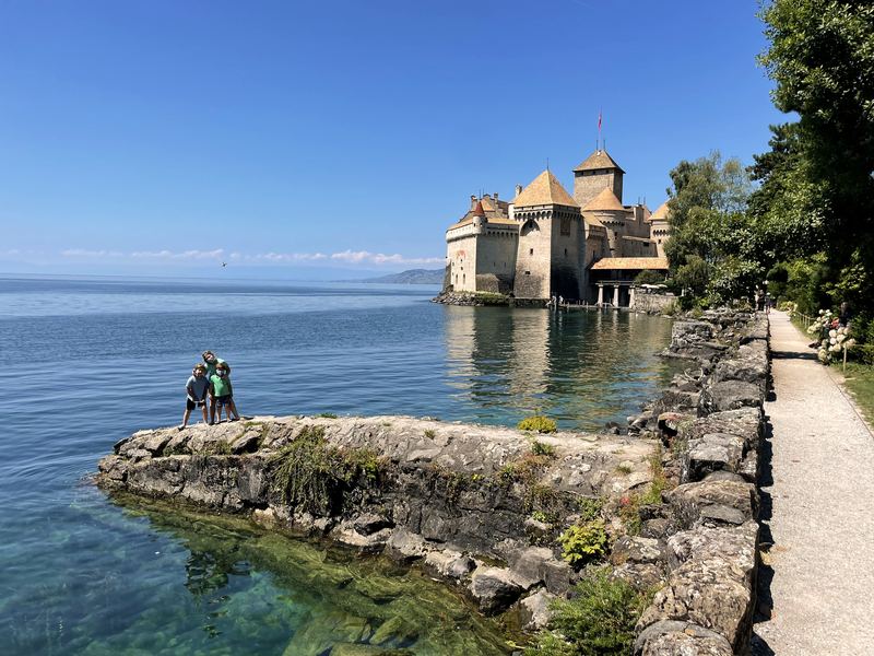Chillon Castle on blue Lake Geneva from a rocky lakeside path.