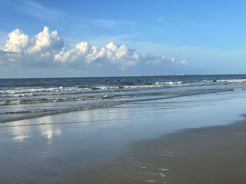 Gentle waves on flat sand under bright blue sky with clouds.