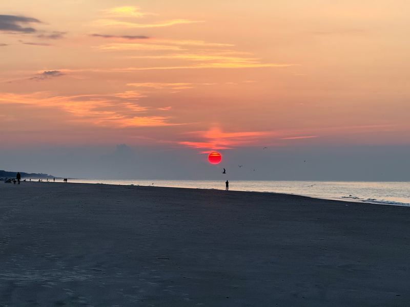 Dark beach at sunset with a small red sun and a distant walker.
